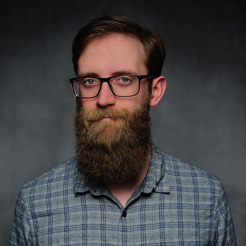 Man with a long beard wearing glasses and a plaid shirt with a dark grey background